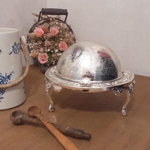 Op de afbeelding: Een zilverkleurige metalen serveerschaal met een koepelvormig deksel voorzien van gedetailleerde bloemmotieven in reliëf. De schaal rust op vier sierlijke, gebogen poten en heeft een brede, decoratieve rand met een glanzende afwerking.