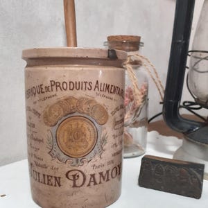 Può includere: Un barattolo cilindrico in ceramica beige con una grafica vintage che riporta il testo FABRIQUE DE PRODUITS ALIMENTAIRES JULIEN DAMOY PARIS 1891, decorato con illustrazioni di monete d'oro e un emblema centrale, con un manico in legno che sporge.