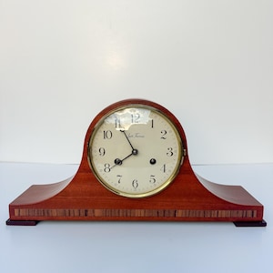 Puede incluir: Un reloj de sobremesa Seth Thomas con caja de madera de caoba pulida, esfera circular de color crema con números arábigos negros, manecillas de metal negro y una franja decorativa incrustada en la base del reloj.