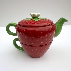 Op de afbeelding: Een keramische theeset in de vorm van een aardbei, bestaande uit een theepot gestapeld op een bijpassend theekopje. De set heeft een glanzende rode afwerking met gele zaaddetails, een groene handgreep en tuit, en een deksel met een witte bloem.