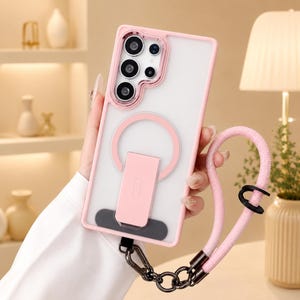 May include: A light pink smartphone case with a frosted back, featuring a circular magnetic ring and a rectangular kickstand. The case includes a matching pink braided wrist strap attached with a dark metal clip and a small black ring connector.