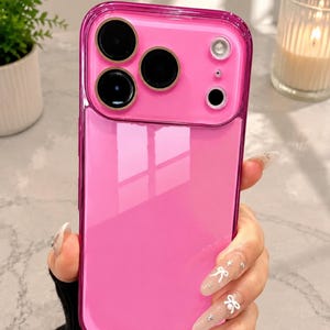 May include: A bright pink smartphone case with a glossy finish, featuring a raised camera cutout section with three circular lens openings and a flash aperture, designed with a clear border frame around the vibrant solid pink back panel.
