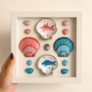 May include: A white square shadow box frame containing a collection of painted seashells. The display features two large scallop shells, one painted pink and one blue, alongside two smaller shells with hand-painted red and blue fish motifs.