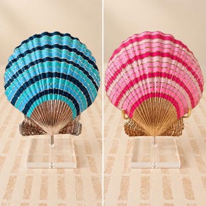 May include: Two decorative scallop shells mounted on clear acrylic stands. One shell features alternating bands of dark blue, light blue, and gold. The other shell features alternating bands of dark pink, light pink, and gold. Both have metallic gold bases.