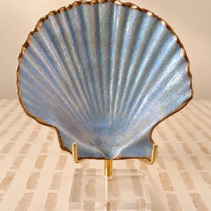 May include: A decorative scallop shell with a shimmering light blue finish and a metallic gold-painted edge. The shell is mounted on a gold-toned metal stand with a clear, square acrylic base, providing a stable display for the iridescent object.
