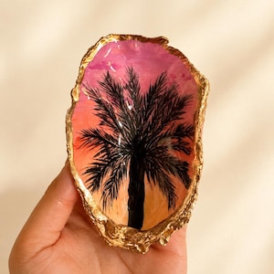 May include: An oyster shell with a hand-painted sunset scene featuring a black palm tree silhouette against a gradient of pink, orange, and yellow. The irregular edges of the shell are finished with a metallic gold trim, creating a decorative dish.