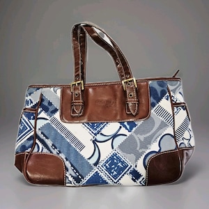 May include: Coach handbag with a patchwork design in shades of blue, white, and gray. The bag features brown leather accents on the handles, trim, and top flap, secured with gold-tone buckles. The Coach logo is visible on the front.