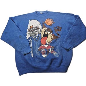 Könnte beinhalten: Blaues Sweatshirt mit einem Cartoon-Bild des Tasmanian Devil beim Basketballspielen. Die Grafik zeigt die Figur mit einem Basketball, einem Korb und einem Netz. Das Sweatshirt hat einen Rundhalsausschnitt und lange Ärmel.
