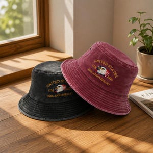 May include: A black and a maroon bucket hat, each featuring an embroidered bald eagle graphic with an American flag, the text UNITED STATES, 1776-2026, and 250th ANNIVERSARY. The hats have a washed, vintage-style fabric texture and stitched brims.