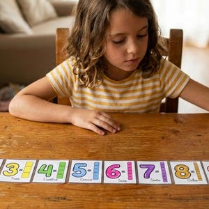 Puede incluir: Un conjunto de diez tarjetas educativas rectangulares dispuestas en una fila horizontal sobre una superficie de madera. Cada tarjeta presenta un número grande y colorido del uno al diez, un bloque de color y el nombre escrito en español.