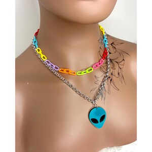 Op de afbeelding: Een gelaagde ketting met een bovenste ketting van veelkleurige plastic schakels in rood, blauw, geel, paars, oranje en neongroen. De onderste ketting is een zilverkleurige metalen schakelketting met een blauwe alien-hoofdhanger met zwarte ogen.