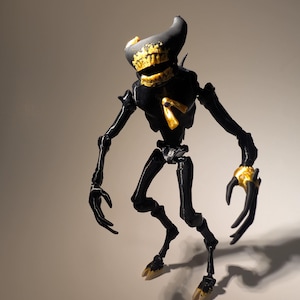 May include: A black articulated action figure with a horned head, featuring gold-colored accents on the face, chest, hands, and feet. The figure has a slender, skeletal build with long limbs, sharp claws, and a small purple detail on the chest piece.