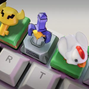 Puede incluir: Tres teclas personalizadas para teclado mecánico con figuras en miniatura: una llave amarilla sobre una base verde, una espada morada en una base de piedra gris y un pollo blanco con cresta roja sobre una base verde, en un teclado blanco.