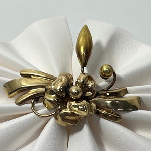 May include: A gold-toned flower brooch with a textured finish. The brooch features a central flower with multiple petals and a single leaf extending upwards.