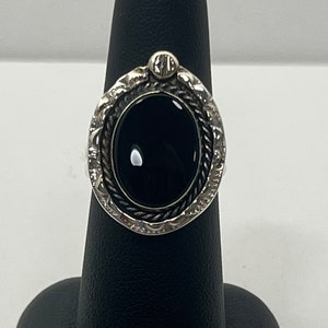 May include: A silver ring with an oval black onyx stone set in a detailed silver bezel. The ring has a textured band and a small silver accent on the top.
