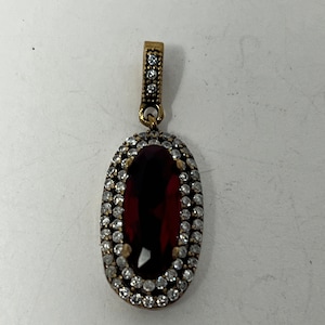 May include: Oval pendant with a large, faceted, dark red gemstone. The gemstone is surrounded by a double row of small, clear crystals. The pendant has a gold-colored metal setting and a small bail with additional clear crystals.