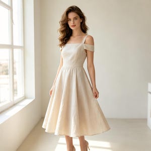 Può includere: Un abito beige di lunghezza midi con scollo quadrato, spalline larghe e maniche scoperte sulle spalle. Il corpetto è aderente e si apre in una gonna svasata e plissettata che arriva sotto le ginocchia, realizzato in un tessuto tinta unita chiaro.
