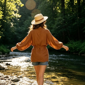 May include: A tan straw hat with a brown band and a long-sleeved, rust-colored linen tunic featuring a belted waist, paired with blue denim shorts. The garment has a loose, flowing fit with gathered sleeves and a straight hemline at the bottom.