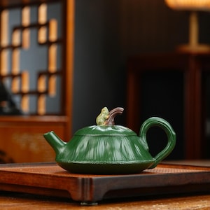 May include: A green ceramic teapot with a frog figurine on the lid. The teapot has a rounded shape with a curved handle and spout. It sits on a wooden tray, showcasing a blend of nature and craftsmanship.
