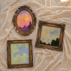 May include: Three ornate gold-toned frames containing landscape art. One oval frame displays a sunset with hot air balloons over purple mountains. Two square frames feature paintings of birds flying across yellow and blue cloudy skies.