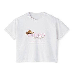 May include: A white short-sleeved t-shirt featuring a brown cowboy hat graphic above the pink text OLIVIA'S and the brown script text Nash Bash, with the date 04.28.2024-04.30.24 printed in a thin brown line box at the bottom of the design.