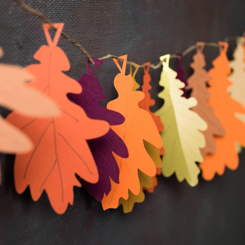May include: A garland of paper oak leaves in various shades of orange, red, yellow, and brown. The leaves are strung together with twine and are hanging against a dark background.