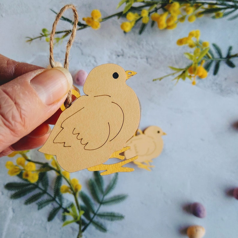 Op de afbeelding: Een gele kuiken-uitsparing met een touwtje eraan, hangend aan een tak van mimosa bloemen. De kuiken is gemaakt van papier en heeft een eenvoudig ontwerp.