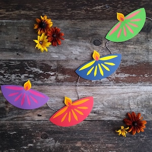 May include: A colorful paper garland featuring diya lamps in shades of purple, red, green, and blue, strung together with twine. The lamps have yellow and orange flame details. The background is a rustic wooden surface with scattered flowers.