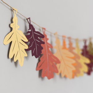 May include: A garland of paper leaves in various shades of brown, orange, and red. The leaves are cut in the shape of oak leaves and are strung together with twine.