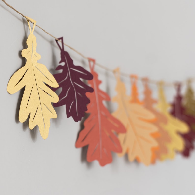 May include: A garland of paper leaves in various shades of brown, orange, and red. The leaves are cut in the shape of oak leaves and are strung together with twine.
