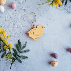 Op de afbeelding: Een lichtbruin houten kuiken ornament met een lus om op te hangen. Het kuiken bevindt zich op een lichtblauwe achtergrond met gele bloemen en gevlekte eieren.