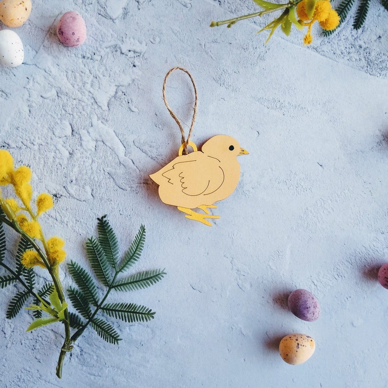 Op de afbeelding: Een lichtbruin houten kuiken ornament met een lus om op te hangen. Het kuiken bevindt zich op een lichtblauwe achtergrond met gele bloemen en gevlekte eieren.