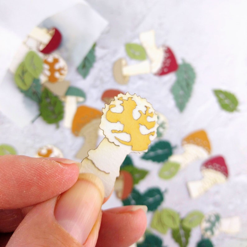 May include: A yellow tree-shaped mushroom charm with a white stem. The charm is made of metal and has a shiny finish. The charm is surrounded by green leaves and other mushroom charms.
