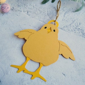Op de afbeelding: Een geel papieren kuiken ornament met een lus van touw om op te hangen. Het kuiken heeft zwarte ogen en een kleine snavel.