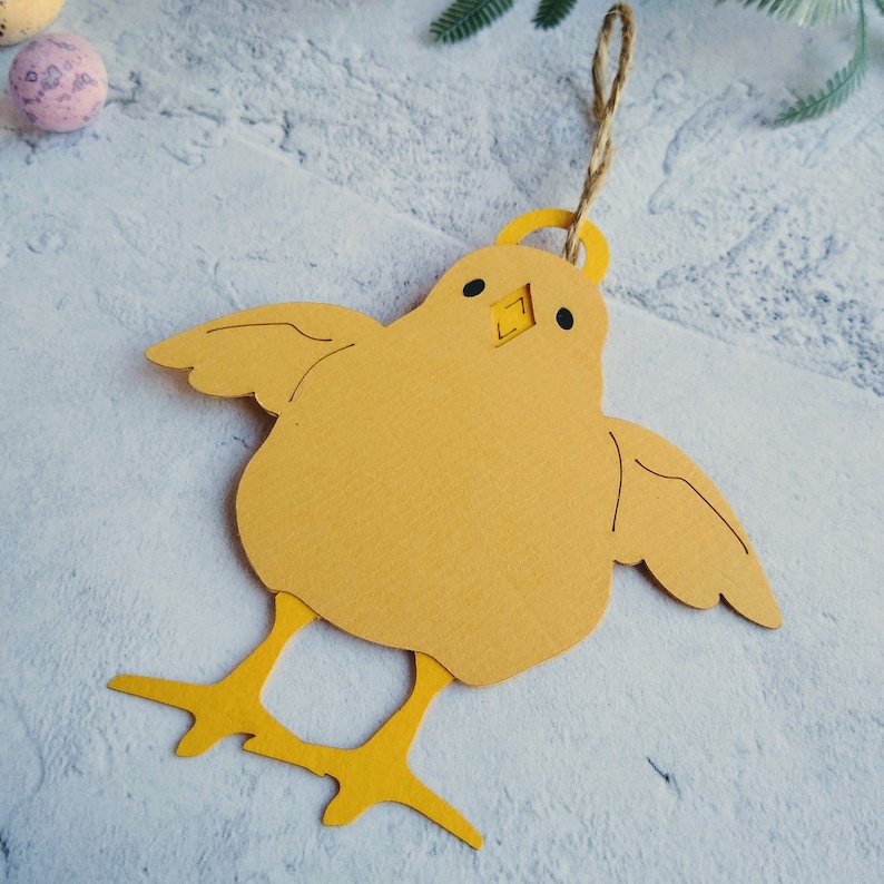 Op de afbeelding: Een geel papieren kuiken ornament met een lus van touw om op te hangen. Het kuiken heeft zwarte ogen en een kleine snavel.