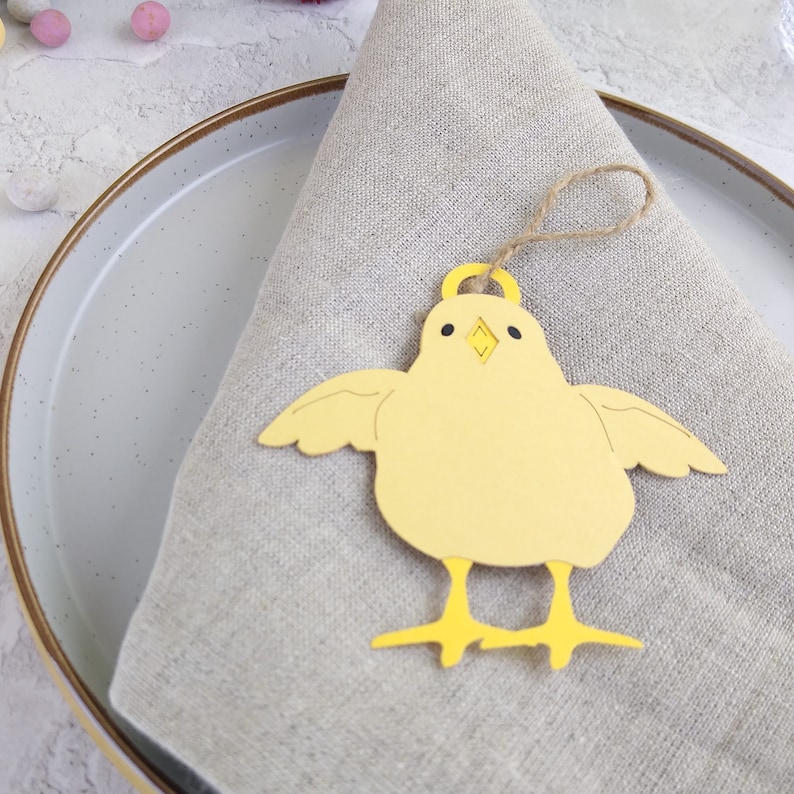 Op de afbeelding: Een gele kuiken plaatskaart met een bruine touwlus. Het kuiken is gemaakt van papier en heeft een eenvoudig ontwerp. De plaatskaart ligt op een beige linnen servet.