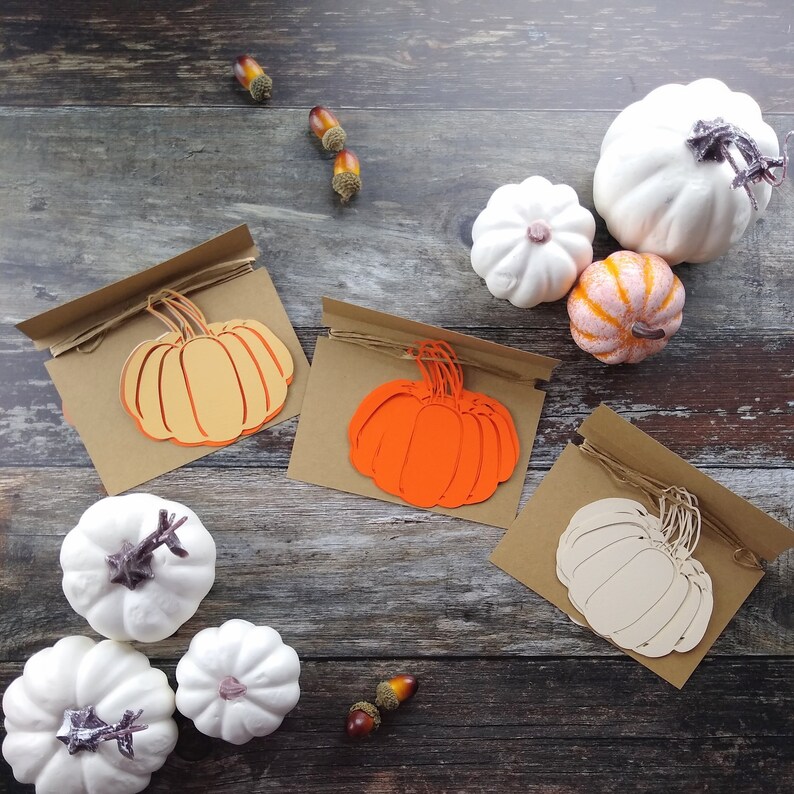 May include: Three brown cardstock cards with a string attached to each. Each card has a different colored paper pumpkin cutout. The pumpkins are orange, peach, and white. The cards are on a wooden surface with white and brown gourds and acorns.