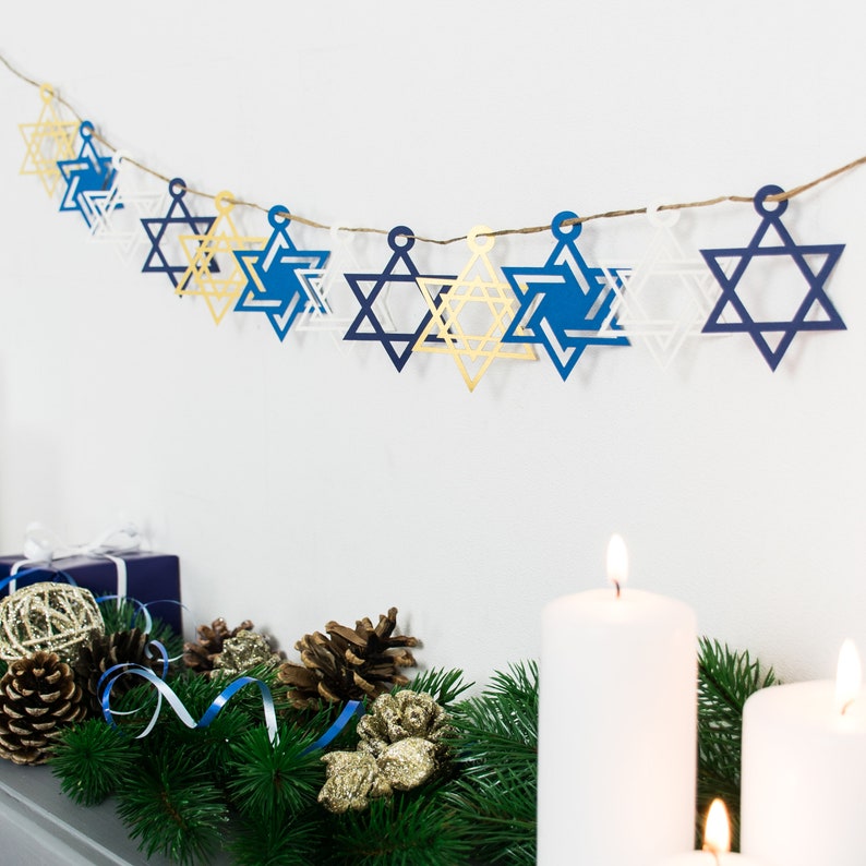 A paper garland with gold,electric blue, white and navy blue star of david paper decorations. Garland is hung on white wall above decorated mantelpiece