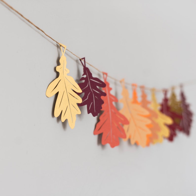 May include: A garland of paper leaves in shades of orange, red, and yellow, strung together with twine. The leaves are cut in the shape of oak leaves.