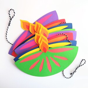 May include: A colorful paper craft resembling a stylized flame or flower. The design features layers of vibrant colors, including green, red, orange, yellow, purple, and blue. A black and white string is attached for hanging.