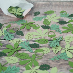 May include: A collection of green paper leaves of various shapes and sizes scattered on a wooden surface. The leaves are cut out of paper and have a delicate, intricate design.