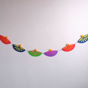May include: A colorful Diwali banner featuring a string of paper lamps in various colors, including purple, red, green, and blue. Each lamp has a yellow flame. The banner is a festive decoration.