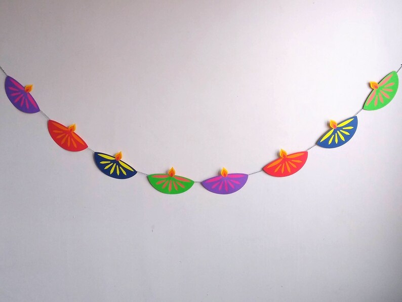 May include: A colorful Diwali banner featuring a string of paper lamps in various colors, including purple, red, green, and blue. Each lamp has a yellow flame. The banner is a festive decoration.