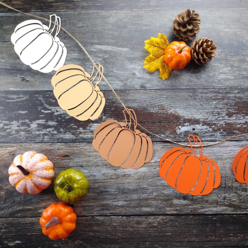May include: A string of paper pumpkins in various shades of orange, white, and brown, with a twine string connecting them. The pumpkins are hanging against a rustic wooden background. There are also small real pumpkins in various colors on the wooden surface.