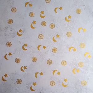 May include: Gold-colored star, crescent moon, and snowflake-shaped confetti scattered on a textured white surface. The celestial-themed decorations are arranged in a random pattern, creating a festive and decorative effect.