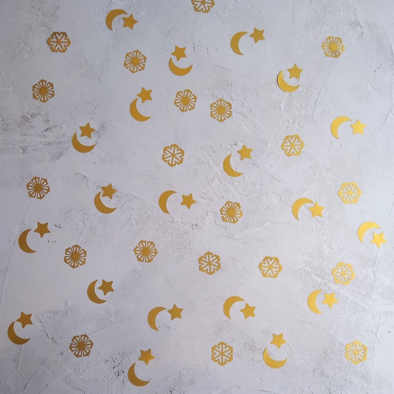 May include: Gold-colored star, crescent moon, and snowflake-shaped confetti scattered on a textured white surface. The celestial-themed decorations are arranged in a random pattern, creating a festive and decorative effect.