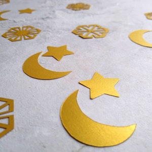 May include: A collection of gold-colored paper cutouts featuring various shapes including crescent moons, five-pointed stars, and geometric snowflake-like patterns scattered across a light gray textured surface.