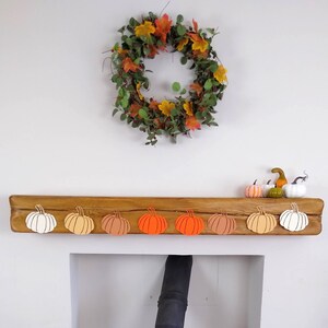 May include: A wooden mantelpiece with a fall-themed wreath above it. The mantelpiece is decorated with a row of eight paper pumpkins in shades of orange, peach, and white.