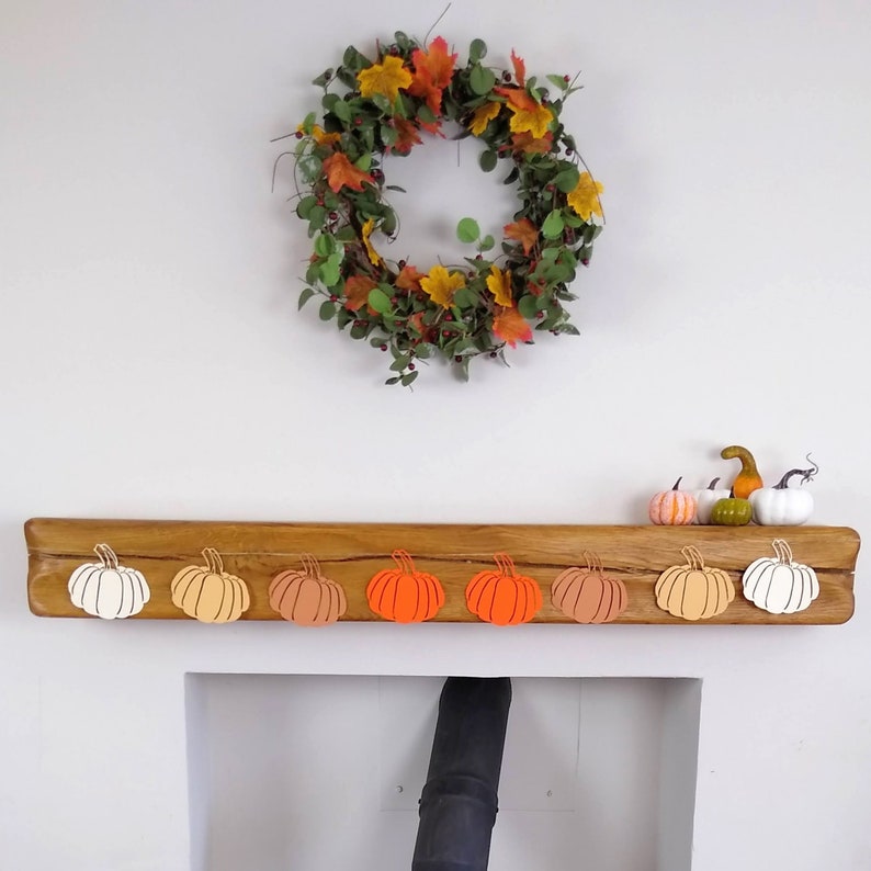 May include: A wooden mantelpiece with a fall-themed wreath above it. The mantelpiece is decorated with a row of eight paper pumpkins in shades of orange, peach, and white.