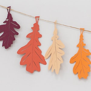 May include: A garland of paper leaves in shades of burgundy, orange, and cream. The leaves are cut in the shape of oak leaves and are strung on a twine string.